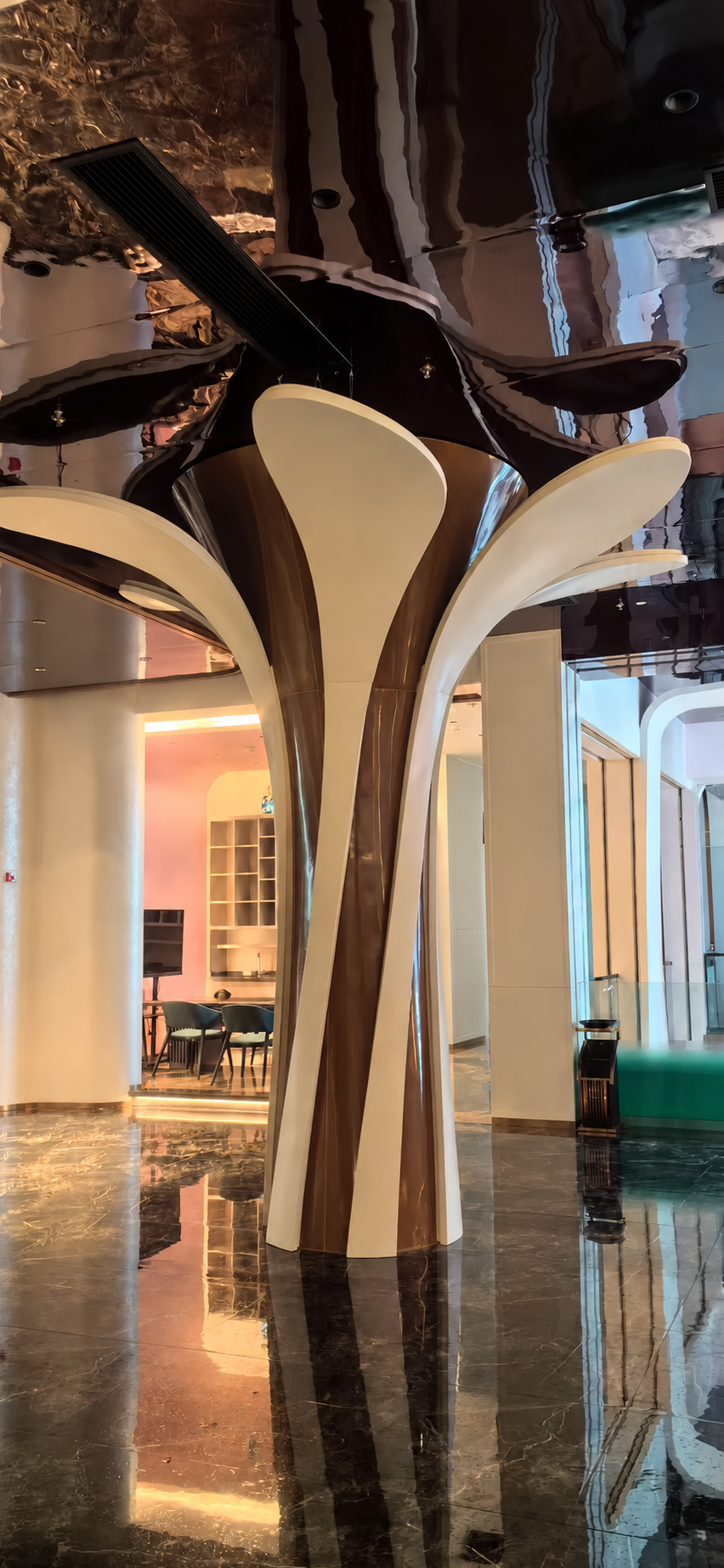 Cream and bronze sculptural tree-form column beneath a mirrored ceiling in an empty lobby with polished marble flooring