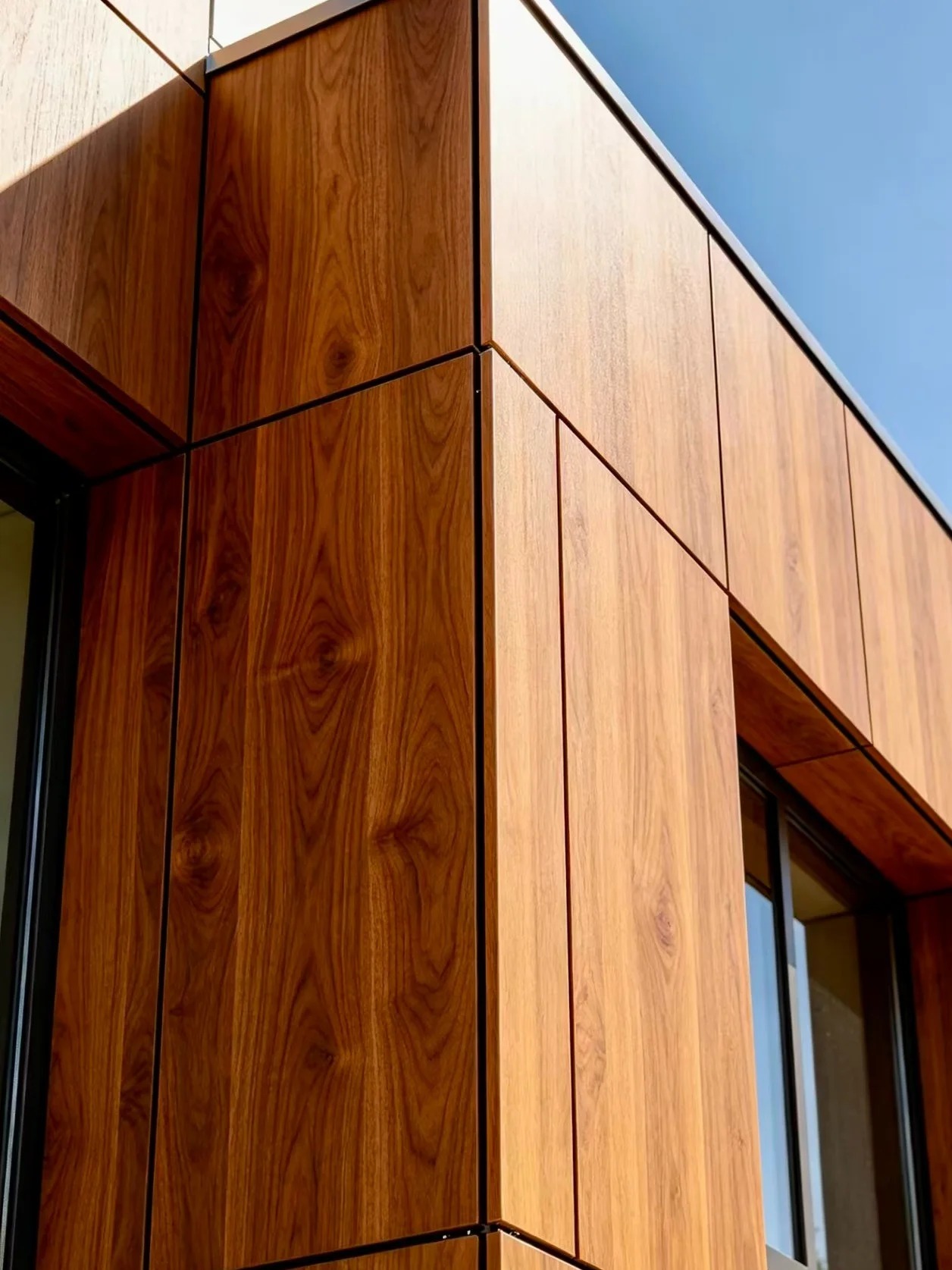 Wood-grain exterior cladding panels meeting at a building corner with dark joints beside a window